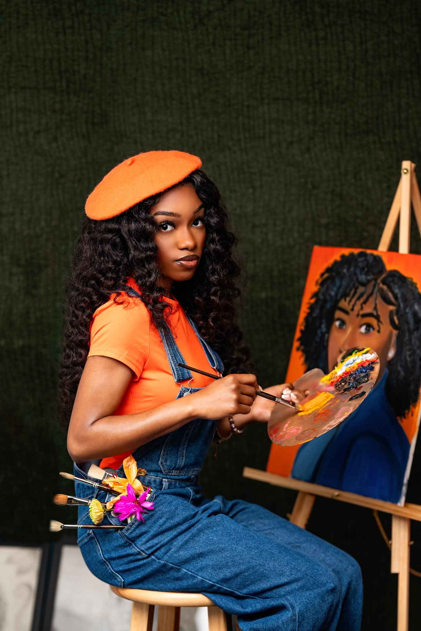 Carlynn in an orange beret and denim overalls seated beside her easel